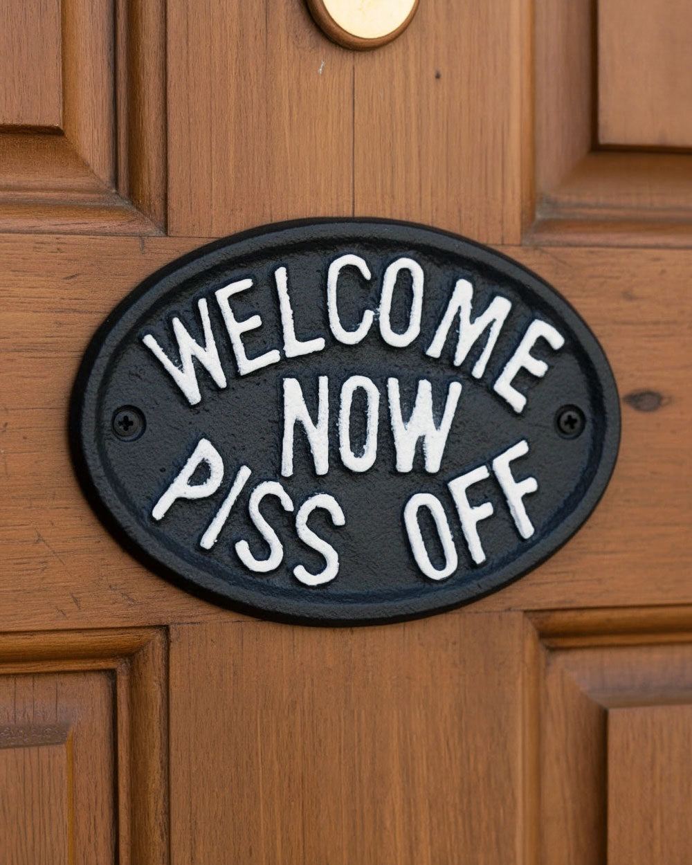 Black oval welcome now sign mounted on wooden door with two screws and white lettering