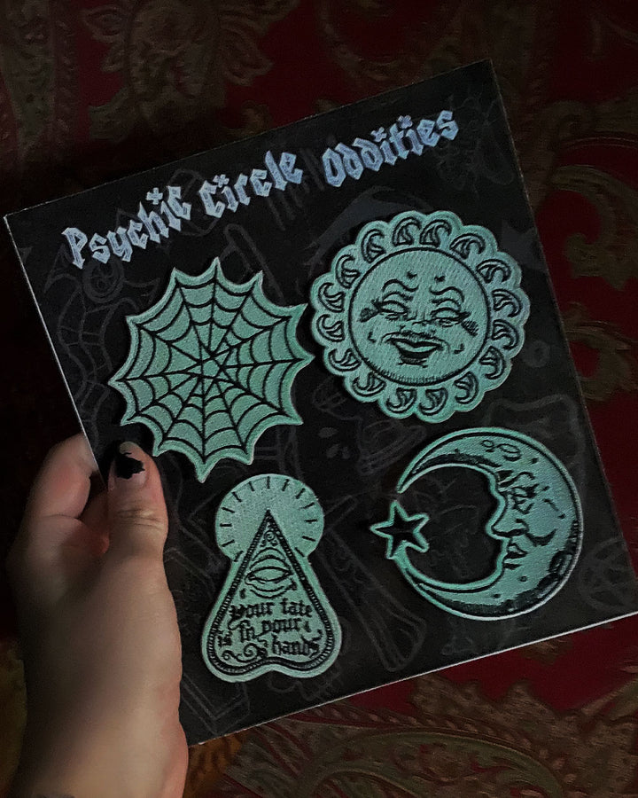 Four glow-in-the-dark patches on a card, showing a sun, crescent moon, spiderweb, and planchette.