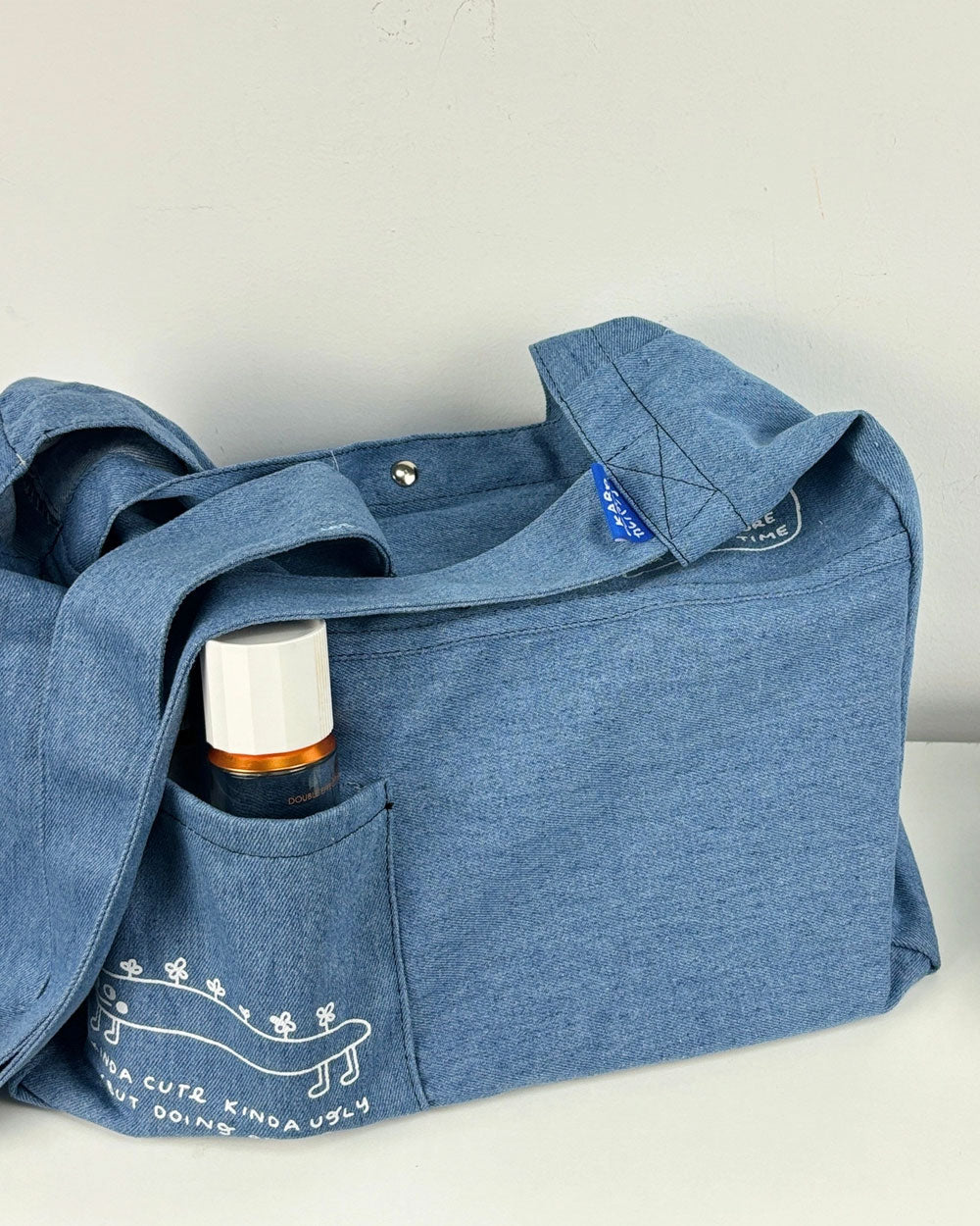 Blue denim tote bag with side pocket and top snap closure, holding a small bottle