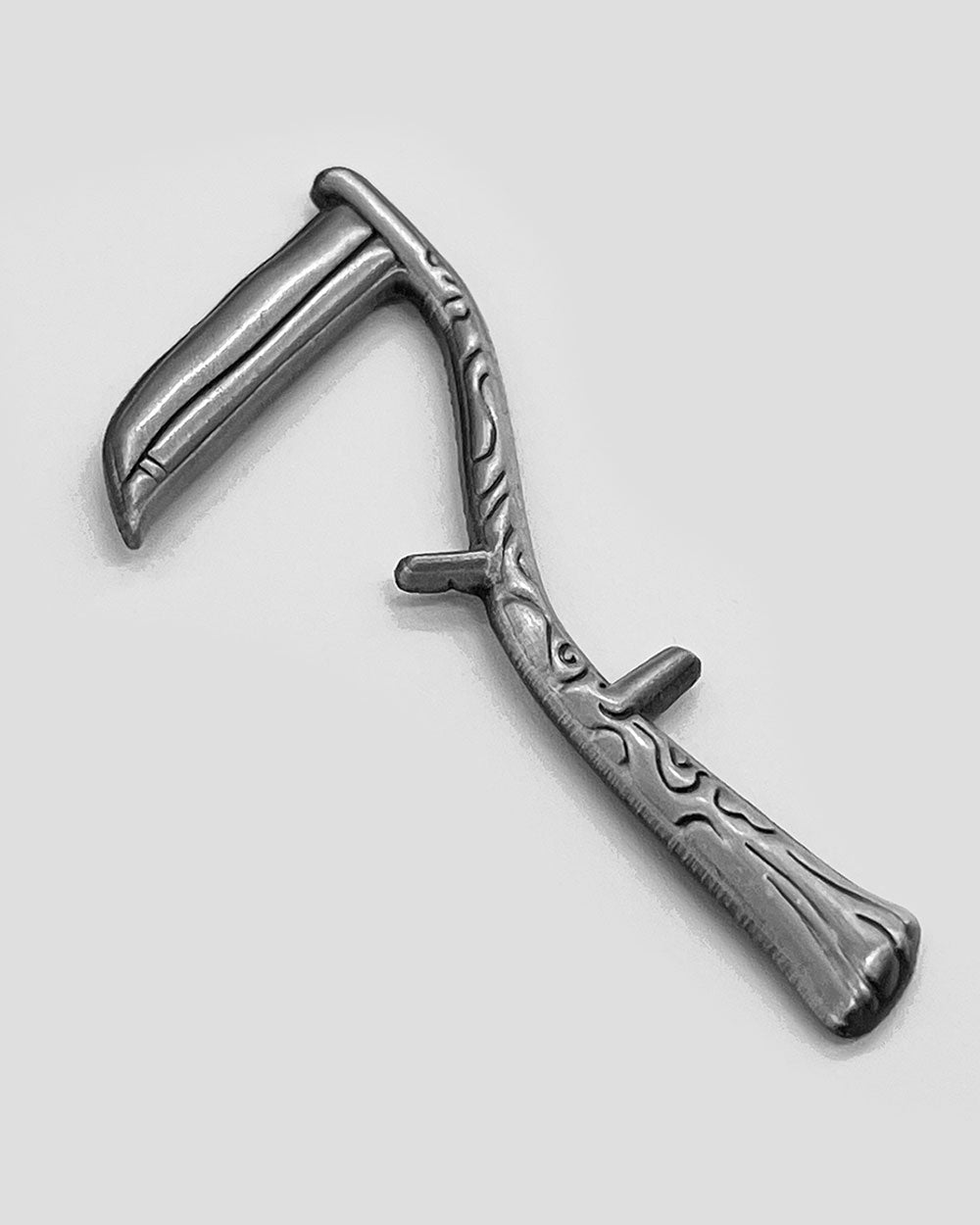 Silver-tone scythe lapel pin, angled, with engraved grip and raised blade